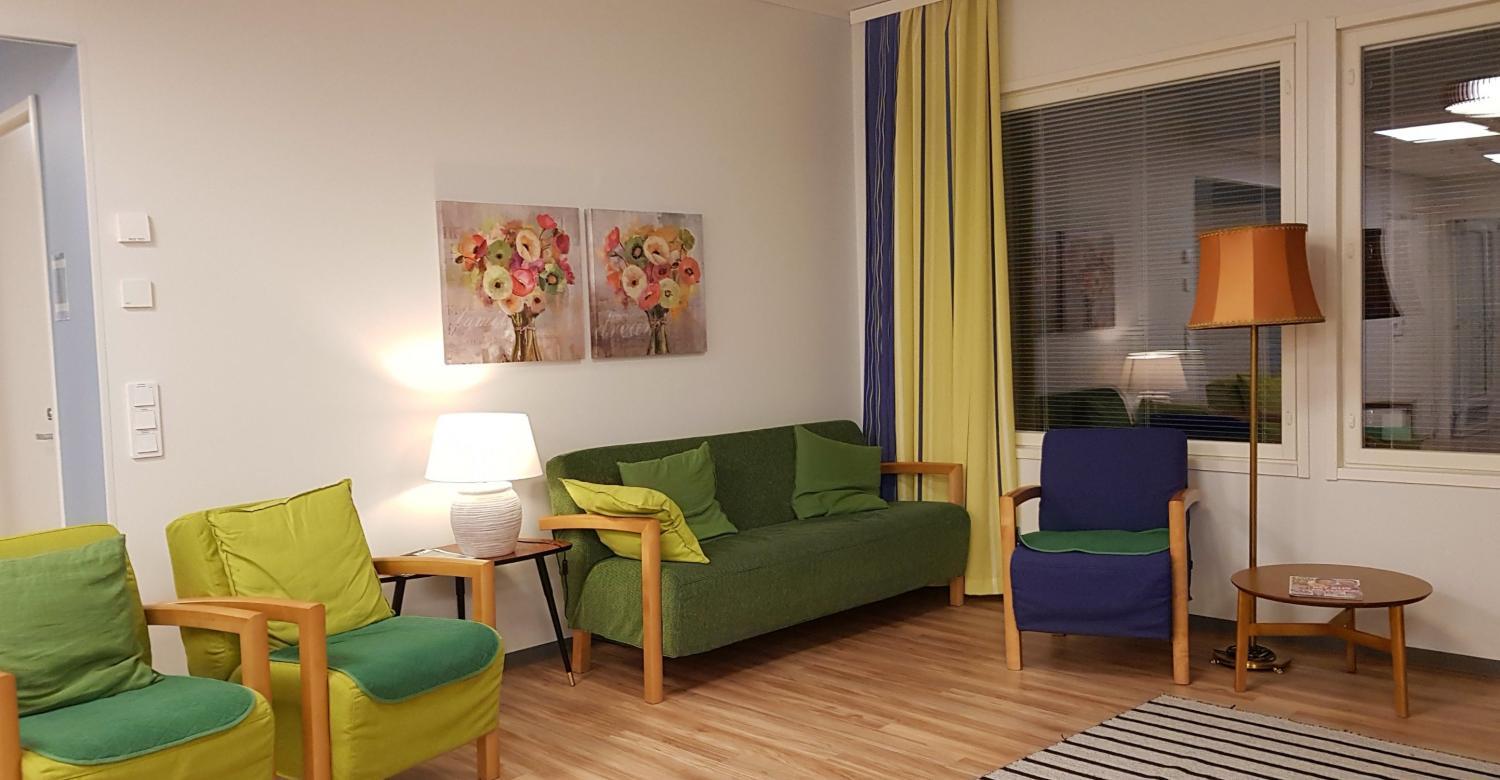 Ett vardagsrum med två gröna fåtöljer, en grön soffa, en blå och grön fåtölj, en golvlampa, ett sidobord med en vit lampa, blomsterkonst på väggen och randiga gardiner bredvid stora fönster.