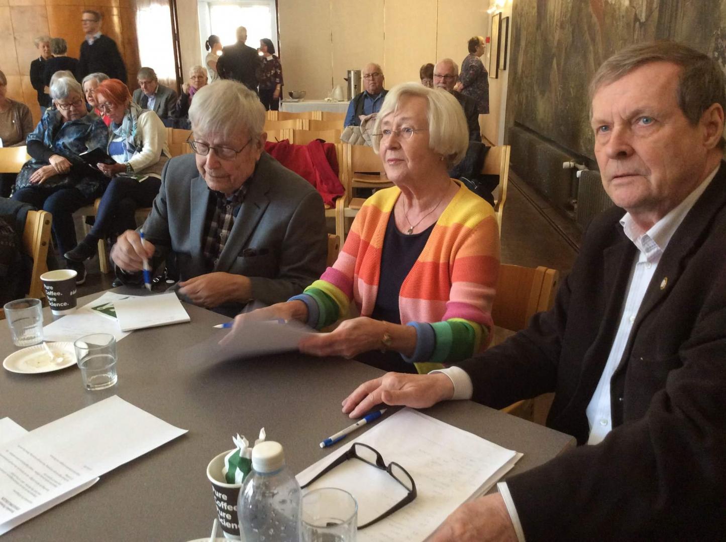 Tre äldre vuxna sitter vid ett bord med papper, pennor och drycker, vända framåt. Fler personer sitter i bakgrunden, några pratar. Miljön verkar vara ett möte eller evenemang i ett gemensamt utrymme.