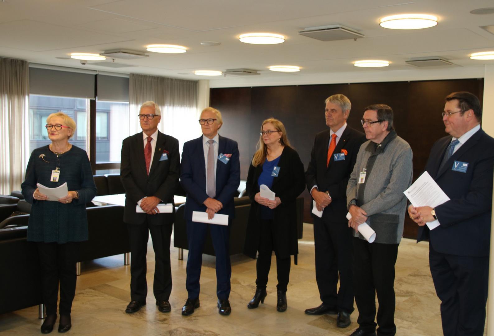 Sju personer klädda i affärskläder står inomhus i ett väl upplyst rum, med papper i handen och namnskyltar. De ser ut att delta i ett formellt möte eller evenemang.