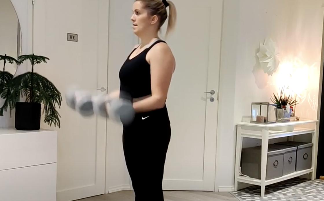 En person i svarta sportkläder står inomhus och håller och lyfter två hantlar med båda armarna böjda vid armbågarna. Rummet har vita väggar, en krukväxt och ett bord med inredningsföremål.