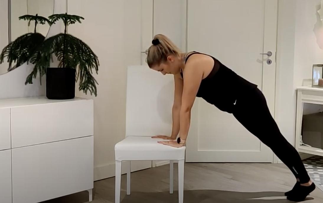 En kvinna i träningskläder utför en lutande armhävning med händerna på en vit stol i ett modernt, minimalistiskt rum.