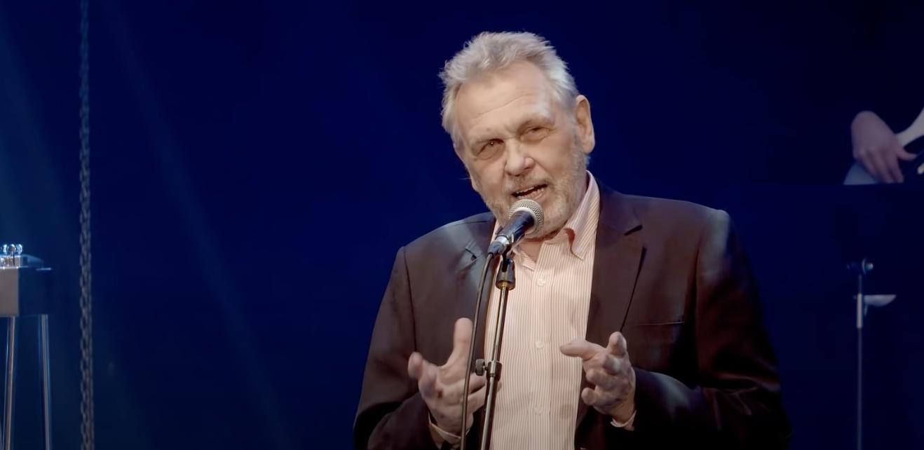 En äldre man i kavaj och ljus skjorta står på scenen och talar i en mikrofon, med blått ljus och suddiga musikinstrument i bakgrunden.