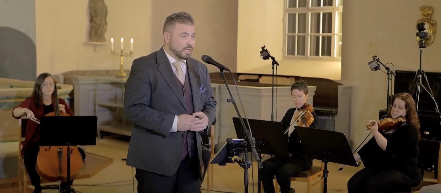 En man i kostym sjunger i en mikrofon i ett väl upplyst rum, ackompanjerad av tre musiker som spelar stråkinstrument och sitter bakom notställ. Teknisk utrustning syns i bakgrunden.