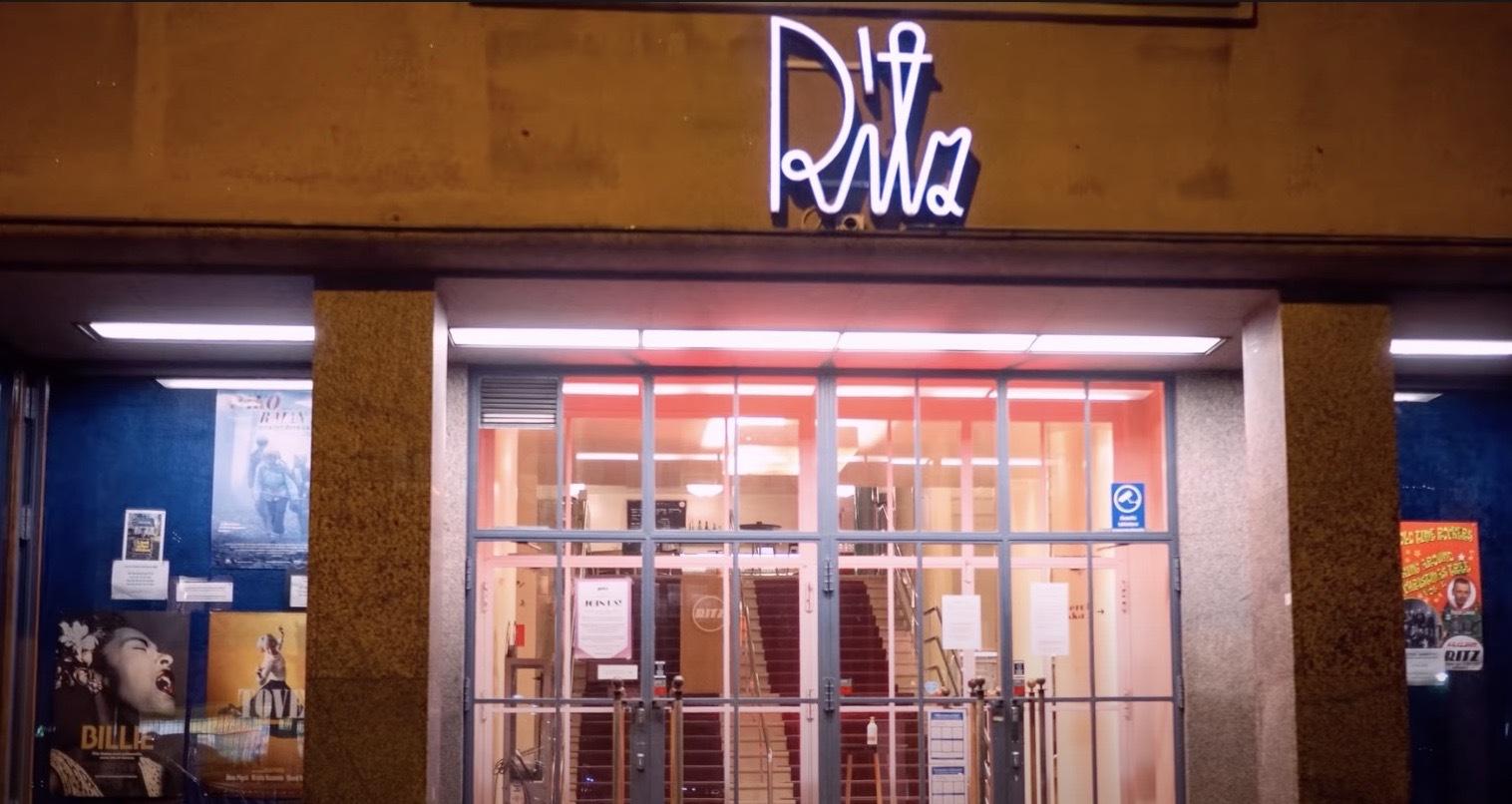 Entrén till en byggnad med glasdörrar och en neonskylt med texten "Riv" ovanför. Affischer hänger på blå väggar på vardera sidan om dörrarna, och en rödbelyst trappa syns inuti.