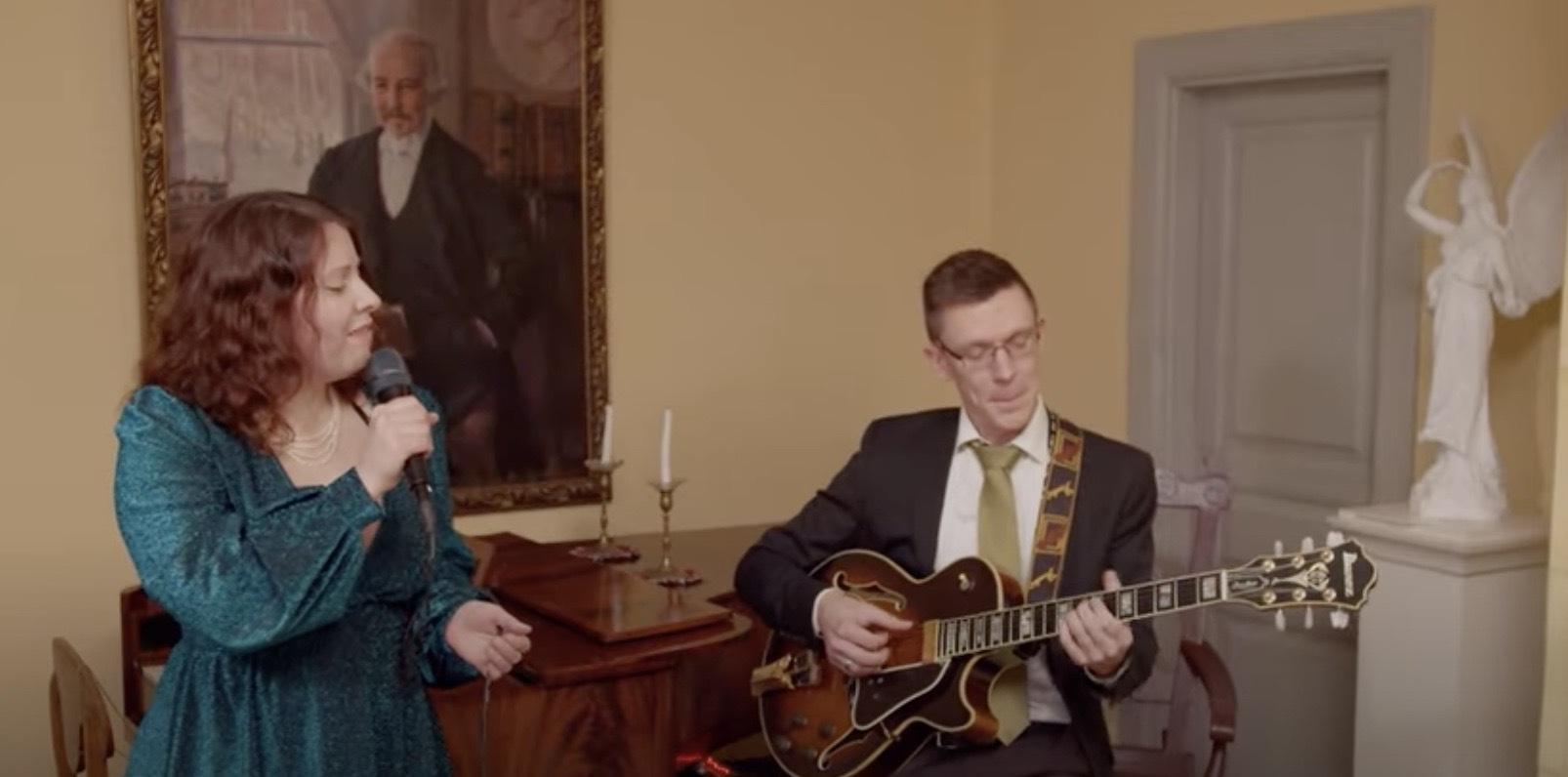 En kvinna i en blågrön klänning sjunger i en mikrofon medan en man i kostym spelar elgitarr. De är inomhus nära ett träpiano, med ett stort porträtt och en vit staty i bakgrunden.