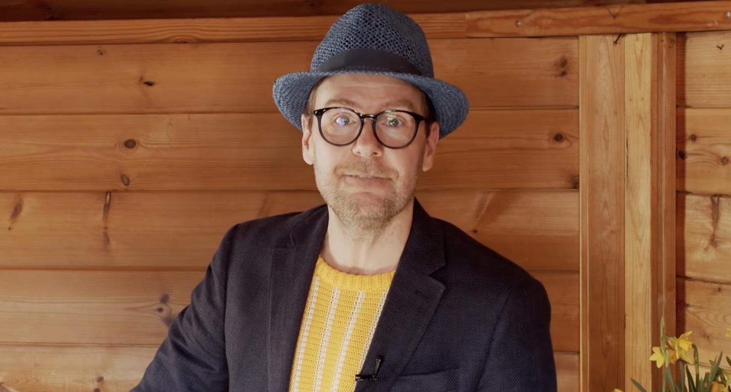 En man med glasögon, blå hatt, mörk kavaj och gul tröja står framför en trävägg och tittar rakt in i kameran.