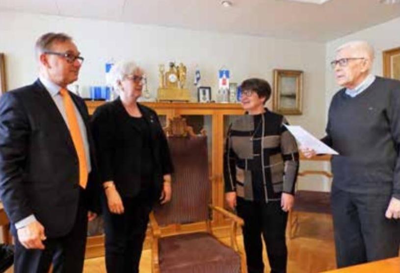 Fyra vuxna står inomhus i ett kontorsliknande rum. En man håller i ett papper medan de andra lyssnar. I bakgrunden finns stolar, en klocka, inramade bilder och diverse föremål på hyllor.