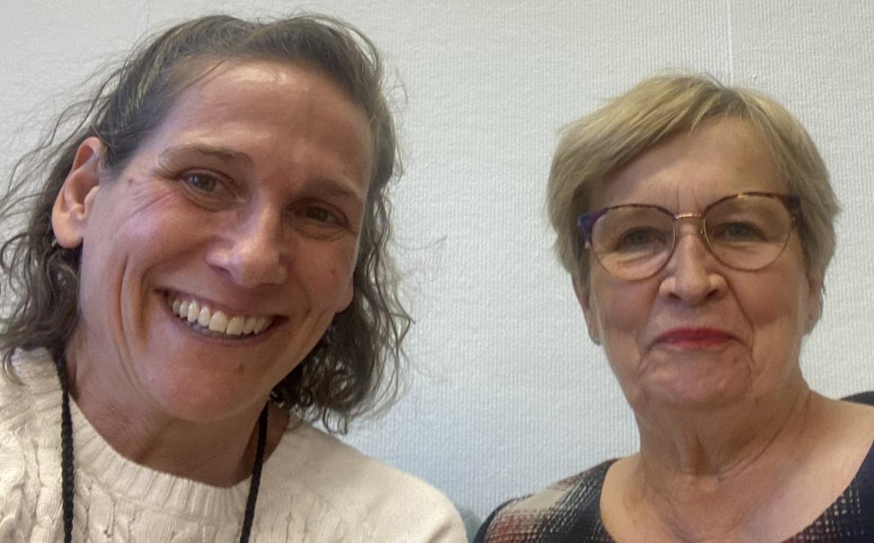Två kvinnor sitter inomhus framför en ljus vägg. Kvinnan till vänster har vågigt brunt hår och ler, medan kvinnan till höger har kort blont hår, glasögon och ett neutralt uttryck.