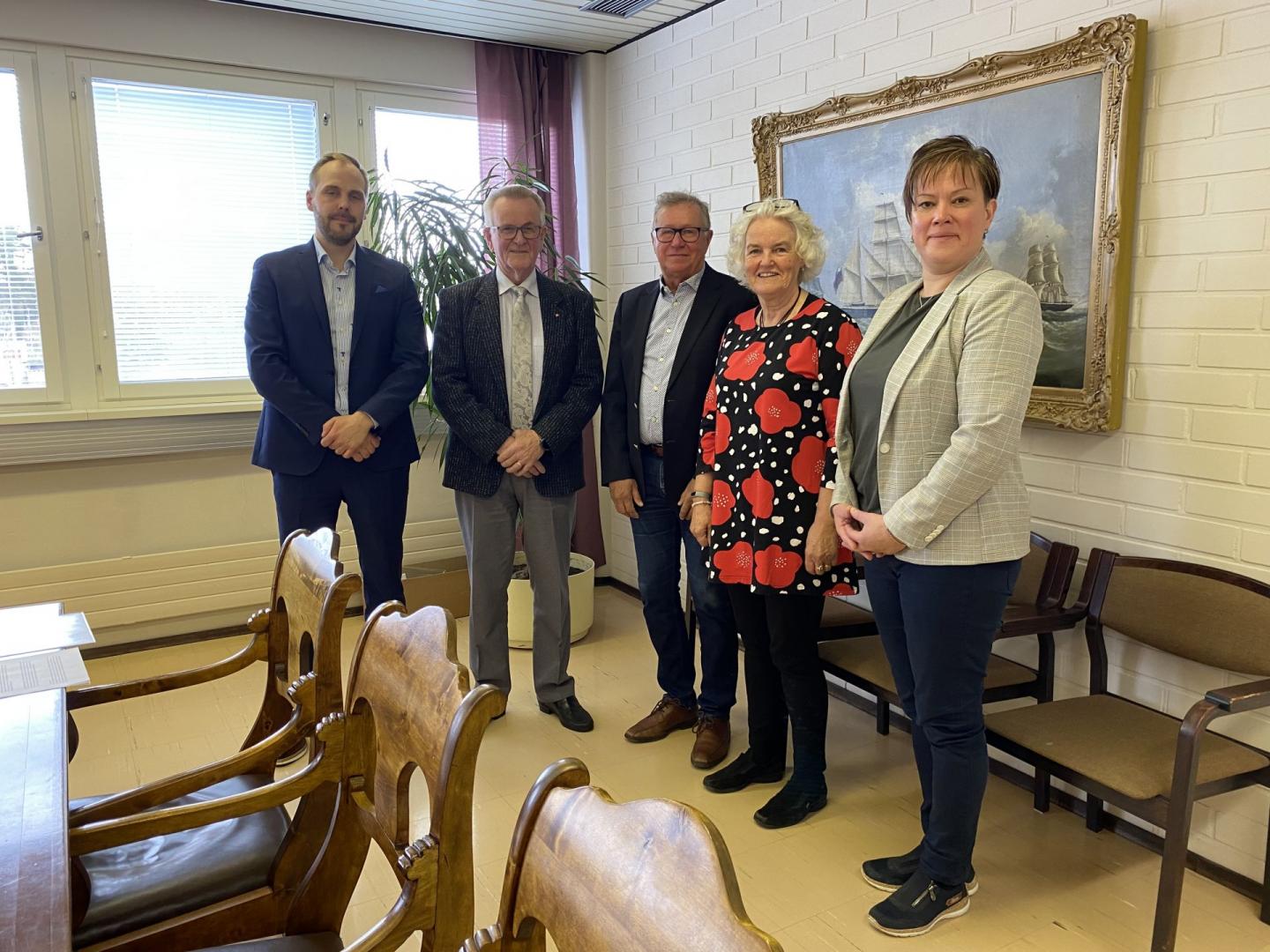 Fem vuxna står sida vid sida i ett mötesrum med trästolar, en stor inramad målning på en vit tegelvägg och fönster som släpper in naturligt ljus. Tre män är i kostym; två kvinnor bär koftor och mönstrade toppar.