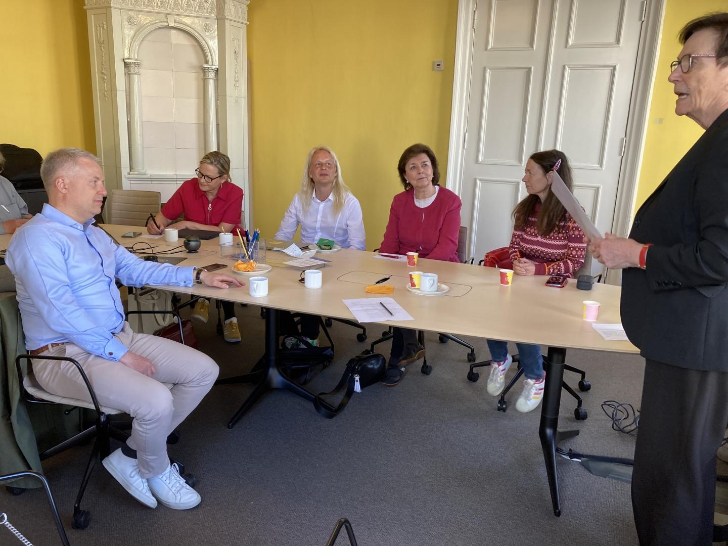 Sex vuxna sitter runt ett mötesbord med papper, kaffekoppar och pennor. En person står, håller i ett dokument och talar till gruppen. Rummet har gula väggar och en dekorativ vit öppen spis.