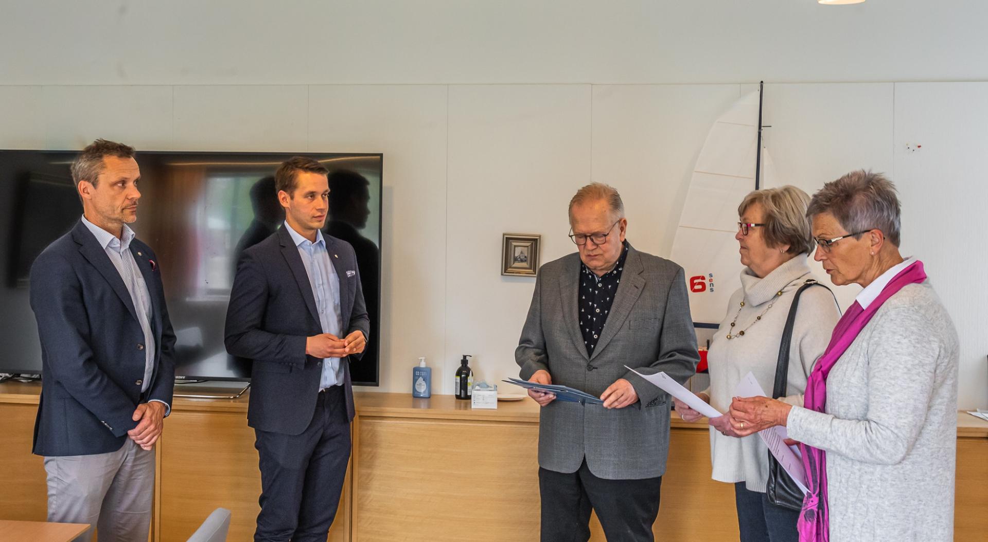 Fem vuxna står inomhus bredvid ett träbord och en TV, håller i papper och diskuterar. Tre män är klädda i kostym, medan två kvinnor bär glasögon och vardagskläder. En modellsegelbåt syns i bakgrunden.