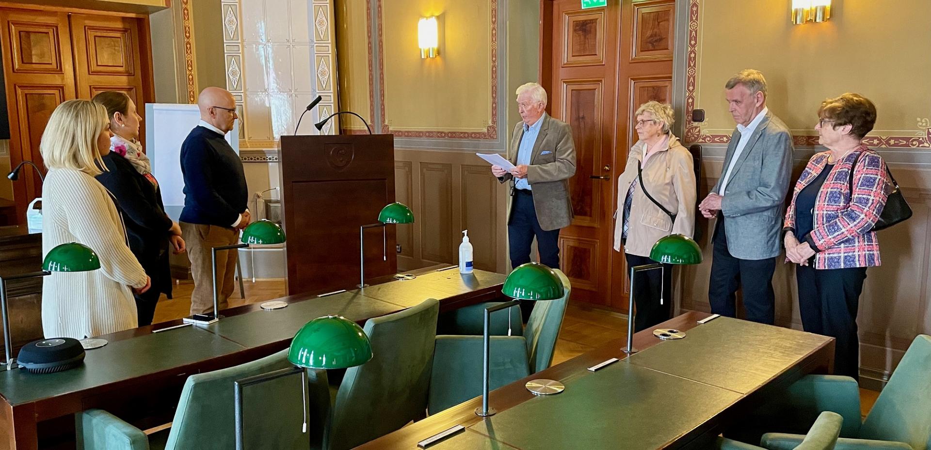 En grupp människor står i ett mötesrum med träpaneler och gröna skrivbordslampor, medan en äldre man vid ett podium läser från en tidning. Sex andra vuxna står mitt emot honom, och en flaska handsprit står på ett skrivbord.