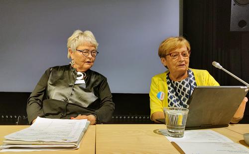 Två äldre kvinnor sitter vid ett bord med papper, ett glas vatten och en surfplatta. En kvinna bär en svart outfit och glasögon, medan den andra bär en gul kofta. De verkar förbereda sig för eller delta i en diskussion.