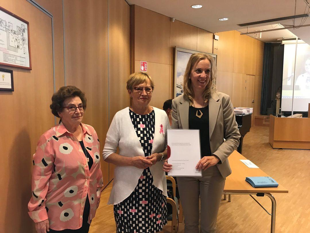 Tre kvinnor står tillsammans i ett konferensrum. En kvinna håller ett dokument och ler. De är klädda i affärskläder. Ett träbord med blå mappar och en projektorduk syns i bakgrunden.