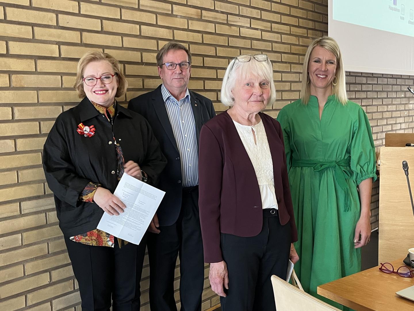 Fyra vuxna står inomhus framför en beige tegelvägg. Tre kvinnor och en man står vänd mot kameran; en kvinna håller i papper och en annan bär en ljusgrön klänning. Ett träpodium syns delvis till höger.