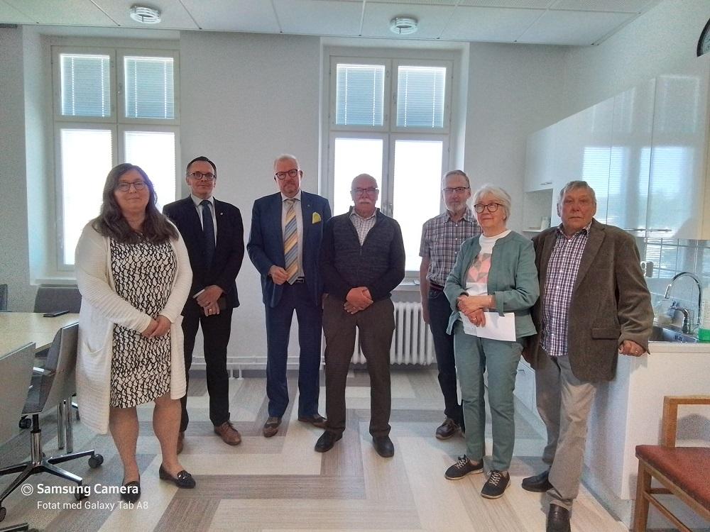 Sju vuxna står i ett ljust rum med stora fönster och ett pentry. De är klädda i affärs- eller halvledig klädsel och poserar för ett gruppfoto. Ett bord och stolar syns till vänster.