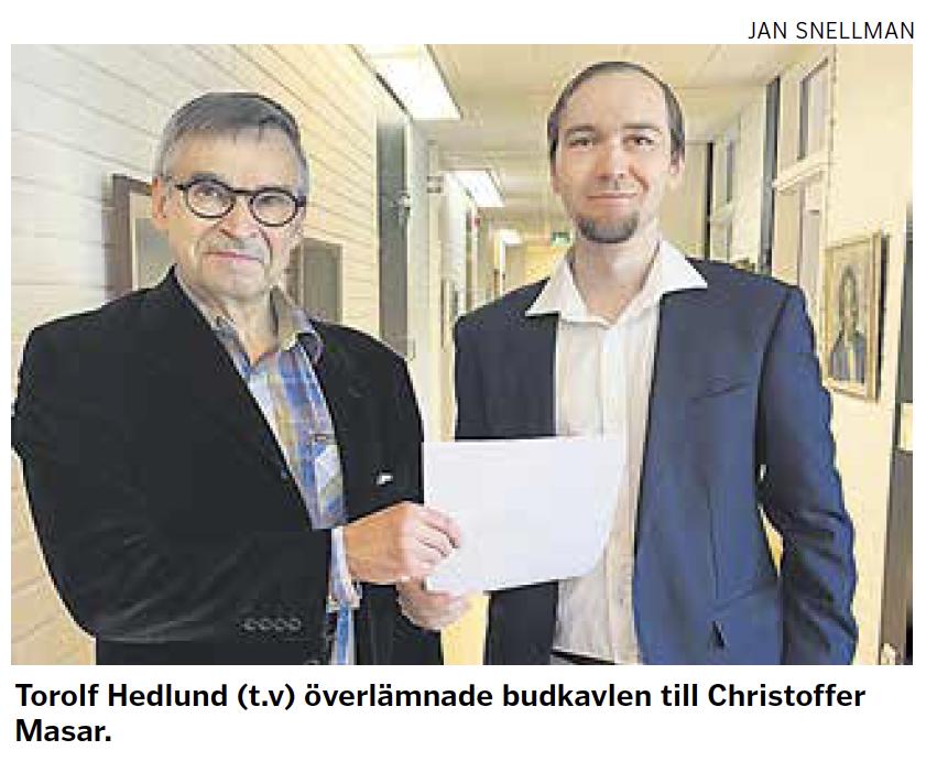 Två män står inomhus nära en hall, en äldre med glasögon och mörk jacka, den andra yngre i ljus skjorta och mörk kostym. Den äldre mannen håller i ett dokument och räcker det till den yngre mannen.