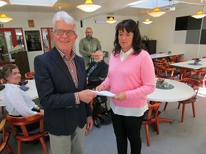 En man i grå kavaj ger ett dokument till en kvinna i rosa tröja. Tre andra personer sitter i bakgrunden i ett rum med runda bord och trästolar.