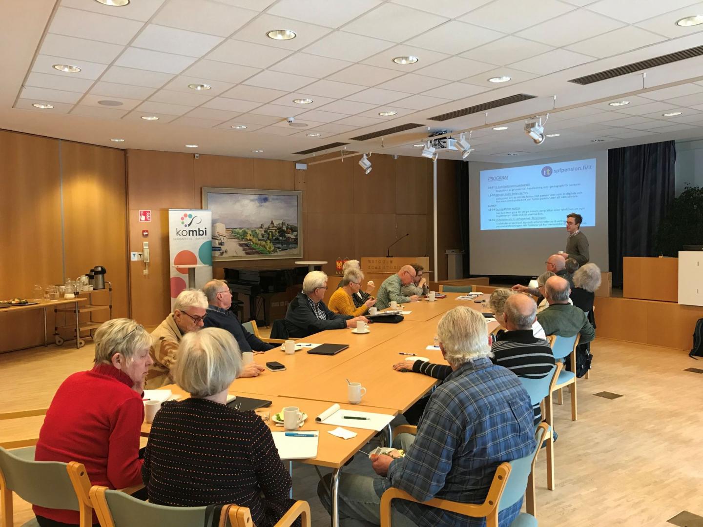En grupp äldre vuxna sitter runt konferensbord mitt emot en presentatör som står nära en projicerad skärm med diabilder. Anteckningsböcker, koppar och papper finns på borden. Rummet har stark belysning och träinredning.
