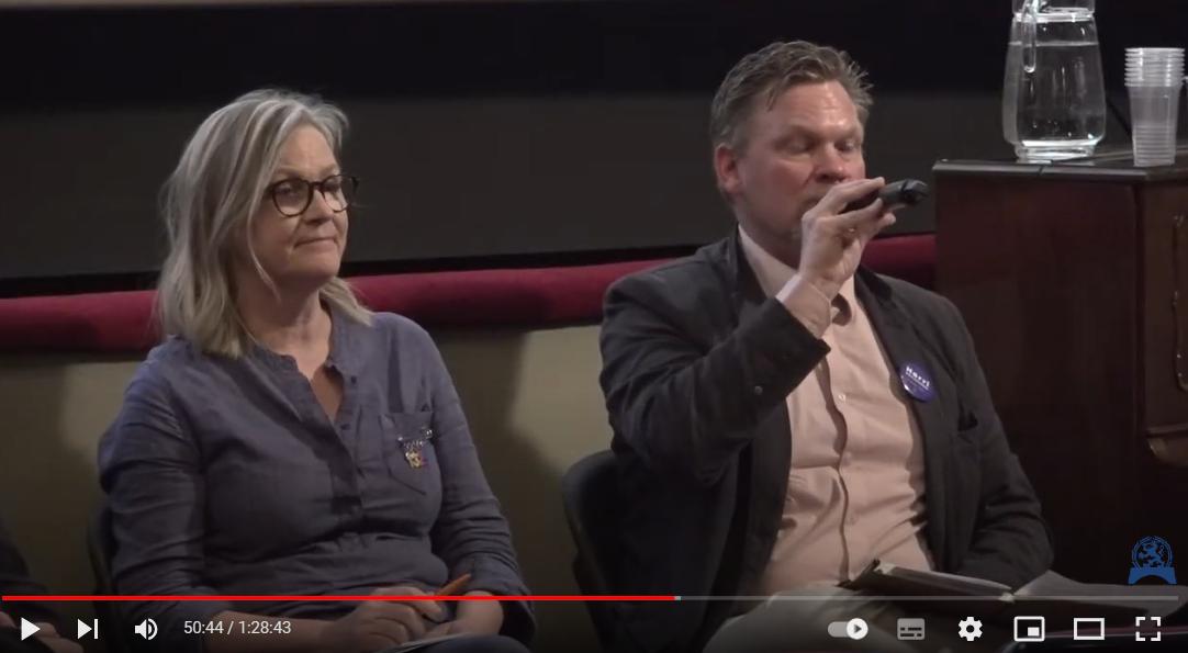 En kvinna med glasögon och blå skjorta sitter bredvid en man i kavaj och ljus skjorta som talar i en mikrofon. De sitter inomhus med en glaskanna med vatten på ett bord bakom sig.