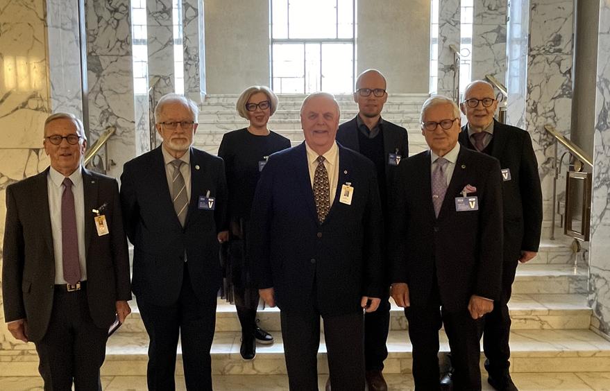 Sju vuxna iklädda formella kläder står på marmortrappor i en byggnad med marmorväggar och stora fönster. Fem män och två kvinnor står vända mot kameran, några med märken. Scenen framstår som professionell och officiell.