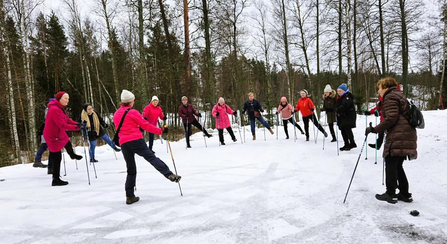 En grupp människor iklädda vinterkläder står i en snöig skogsglänta, håller i vandringsstavar och sträcker ena benet framåt, som om de deltar i en utomhusträning eller ett uppvärmningspass.