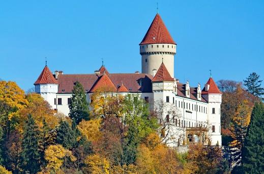 Ett stort vitt slott med röda tak på torn och kanonhuvuden är omgivet av höstträd under en klarblå himmel.