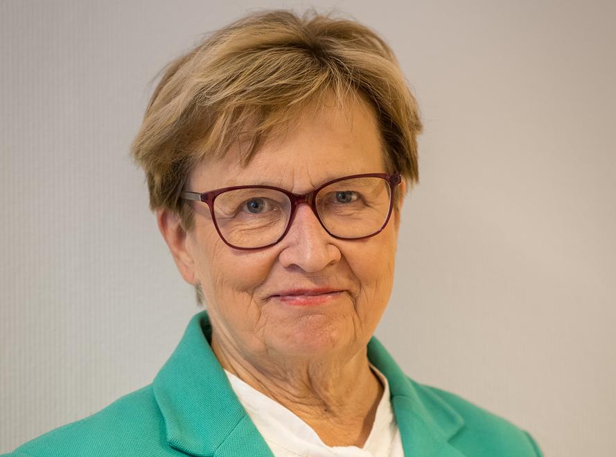 En äldre kvinna med kort ljusbrunt hår och glasögon bär en blågrön kavaj över en vit skjorta och står framför en enfärgad ljus bakgrund.