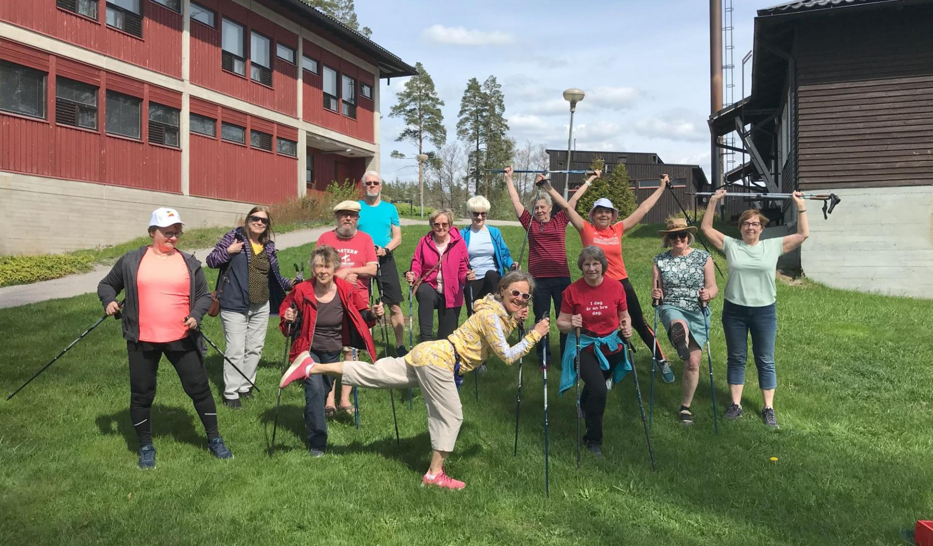 En grupp äldre vuxna står utomhus på gräsmattan, håller i gångstavar, poserar och ler för kameran framför en röd och brun byggnad en solig dag. Några personer har armarna uppe, och en person står på ett ben.