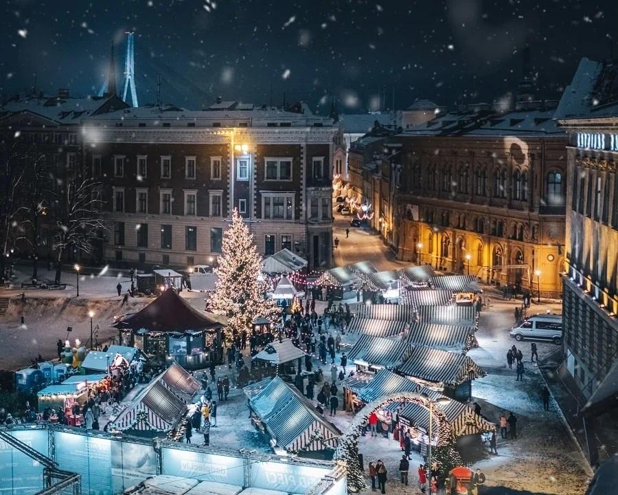 Flygfoto över en festlig julmarknad utomhus på natten, med dekorerade stånd, en stor upplyst julgran, snö på marken och människor som går bland stånden på ett torg omgivet av historiska byggnader.
