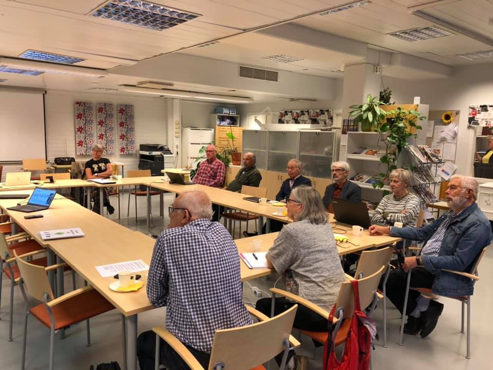 En grupp äldre vuxna sitter runt rektangulära bord i ett väl upplyst mötesrum och lyssnar på en presentation. Bärbara datorer, papper och kaffekoppar står på borden. En whiteboardtavla och en dekorativ vägg syns i bakgrunden.