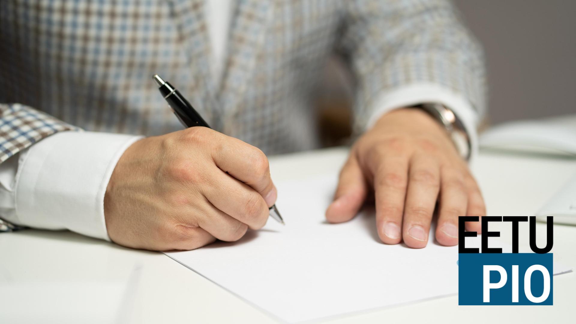 En person i rutig kavaj skriver på ett tomt papper med penna vid ett vitt skrivbord. Bokstäverna EETU PIO visas i det nedre högra hörnet.