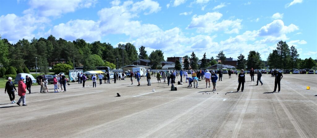 En stor grupp människor står utspridda på en bred, öppen parkeringsplats, några står och andra leker bollar. Flera fordon och träd syns i bakgrunden under en delvis molnig himmel.