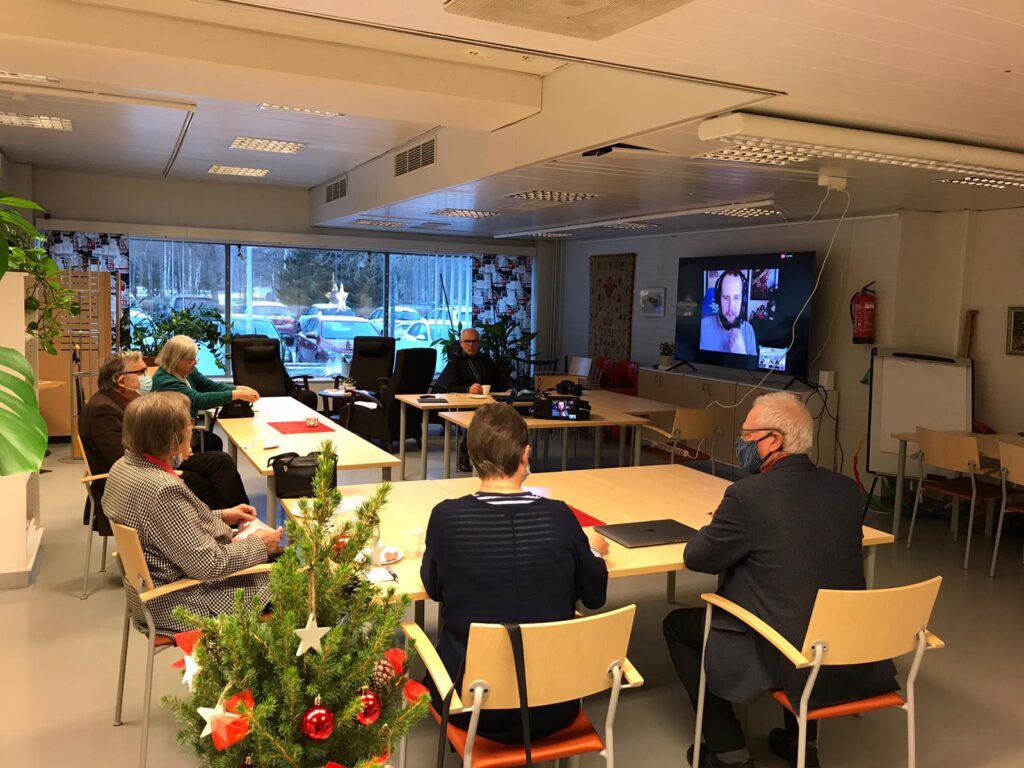 Lagen sitter samlade i hästsko framför en stor skärm med en person som syns i bild med hörlurar på.