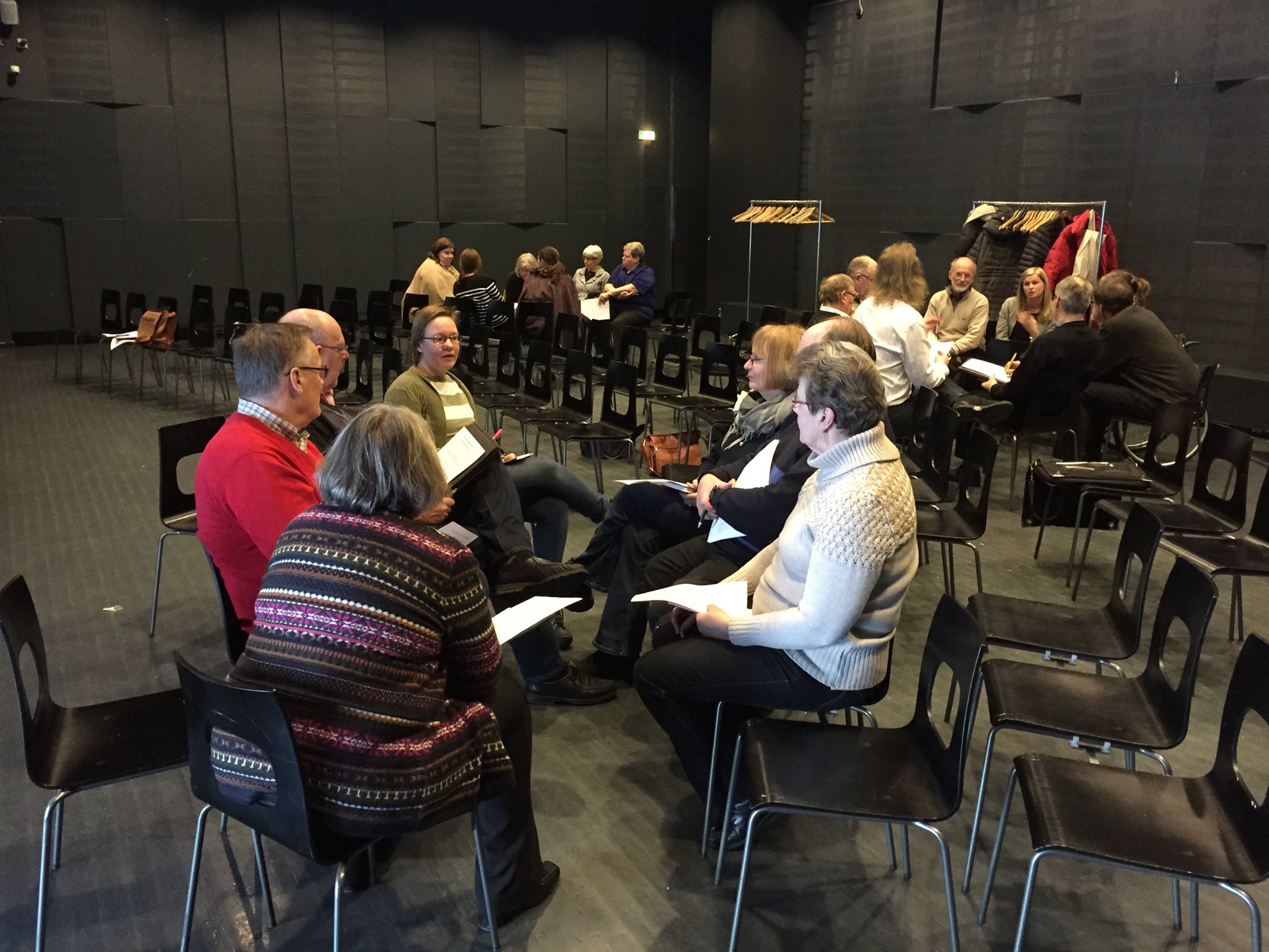 En grupp människor sitter i små cirklar på svarta stolar i ett stort, mörkt rum, engagerade i diskussioner eller gruppaktiviteter. Rockar hänger på hyllor i bakgrunden. Alla verkar fokuserade på samtal eller pappersarbete.