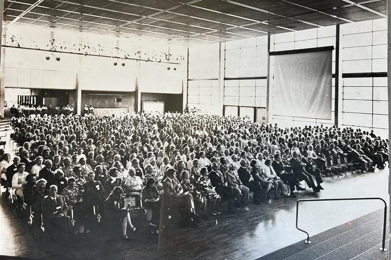 En stor skara människor sitter i rader av stolar i en rymlig, väl upplyst sal med fönster från golv till tak och högt i tak. I förgrunden syns en scen med ett räcke.