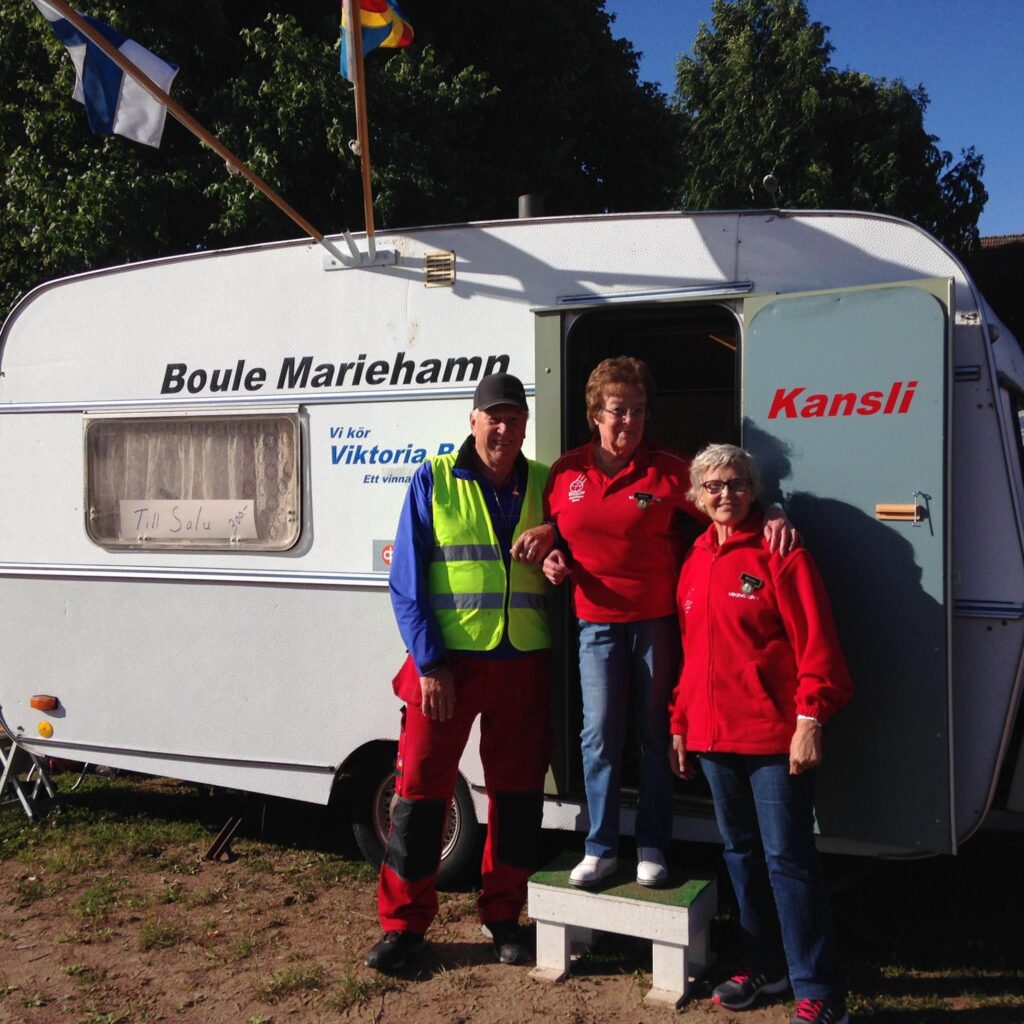 Tre personer står och ler framför en vit husvagn märkt Boule Mariehamn Kansli. De två kvinnorna bär röda toppar och mannen har en neonfärgad säkerhetsväst och blå keps. På en skylt i fönstret står det Till Salu. Träd och flaggor syns ovanför.