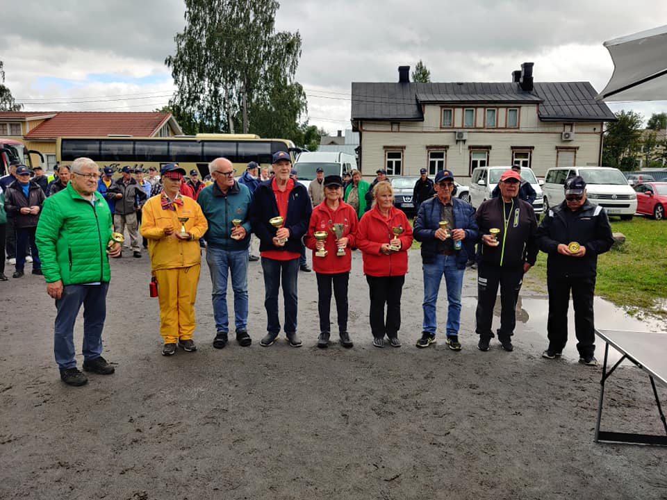 En grupp på tio äldre vuxna, några med pokaler och medaljer, står i en rad utomhus på en grusplan; hus, bilar och fler människor syns i bakgrunden under en molnig himmel.