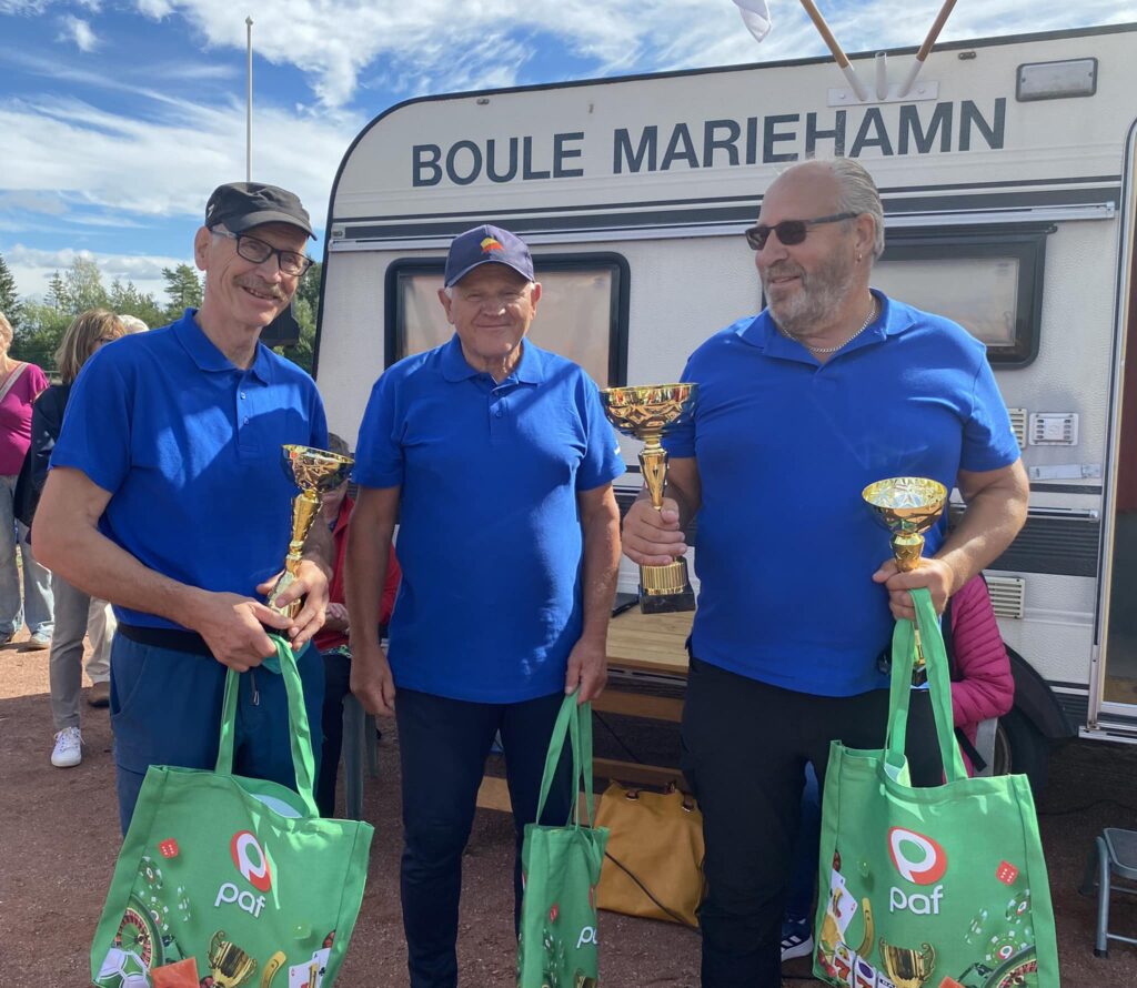 Tre män i blå skjortor står framför en husvagn märkt BOULE MARIEHAMN, var och en håller i en guldtrofé och en grön presentpåse med en paf-logotyp. Det ser ut att vara ett sport- eller tävlingsevenemang.