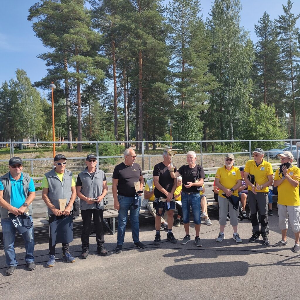 En grupp på nio personer, de flesta sportklädda, står i en kö utomhus en solig dag. Fem personer bär gula skjortor, medan övriga är mörkt klädda. Träd och ett staket syns i bakgrunden.