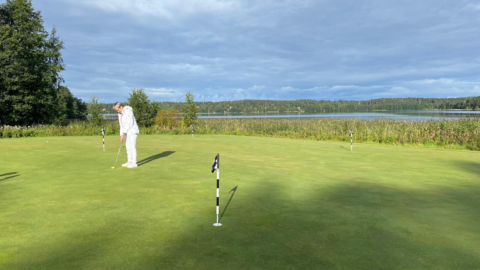 En vitklädd person står på en golfgreen och förbereder sig för att putta. Flera golfflaggor syns, med en sjö och en skogsklädd strandlinje i bakgrunden under en molnig himmel.