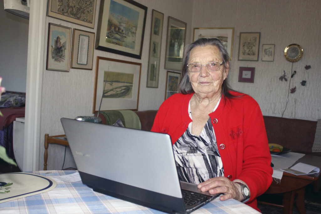 En äldre kvinna med glasögon och röd kofta sitter vid ett bord och använder en bärbar dator. Hon befinner sig inomhus, med inramade bilder och konstverk på väggen bakom henne.