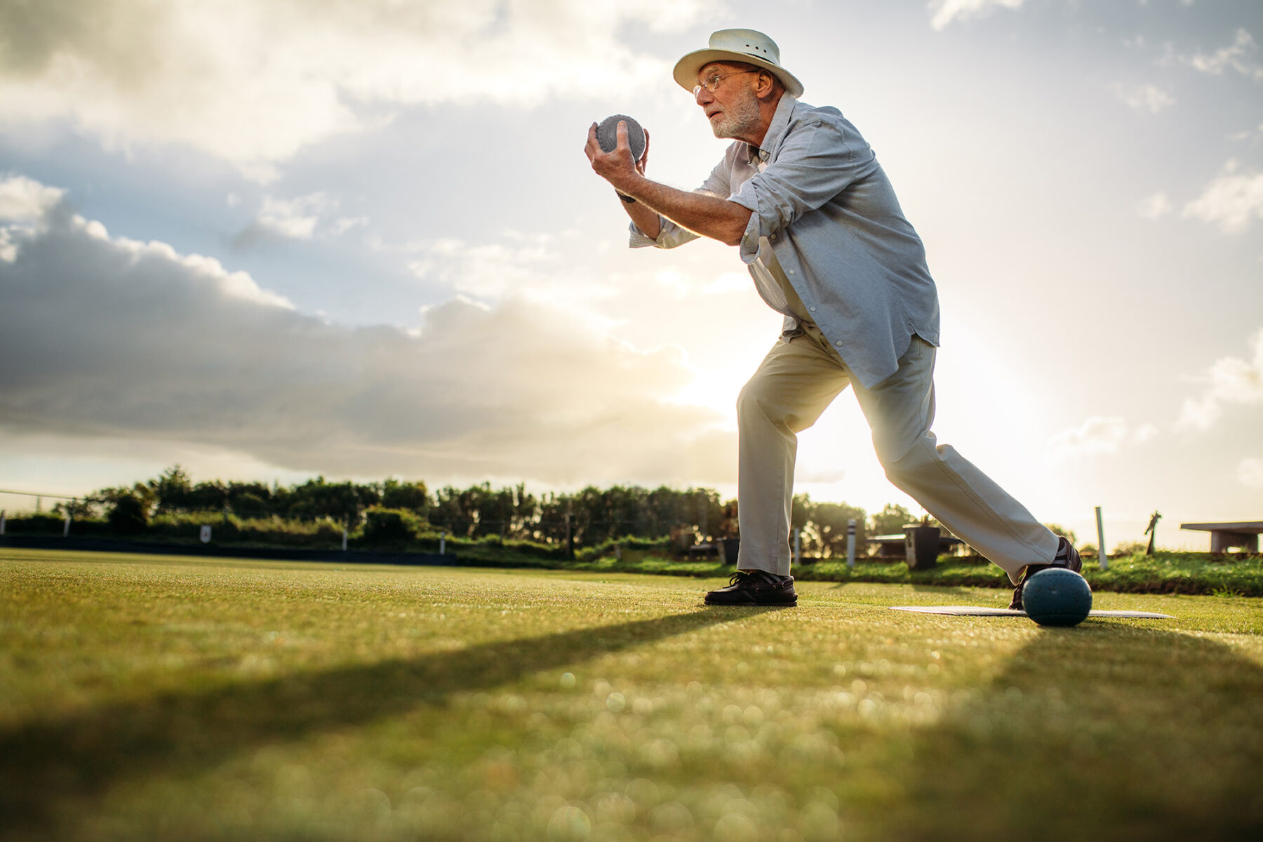 En äldre man i vit hatt och ljusa kläder spelar boule på ett grönt fält i solnedgången, han håller i en skål och förbereder sig för att kasta, med solen lågt på himlen bakom sig.
