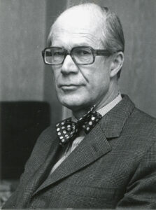 En man med glasögon och kort, prydligt kammat hår bär en prickig fluga och en rutig kostymkavaj och tittar in i kameran med ett neutralt uttryck på ett svartvitt foto.