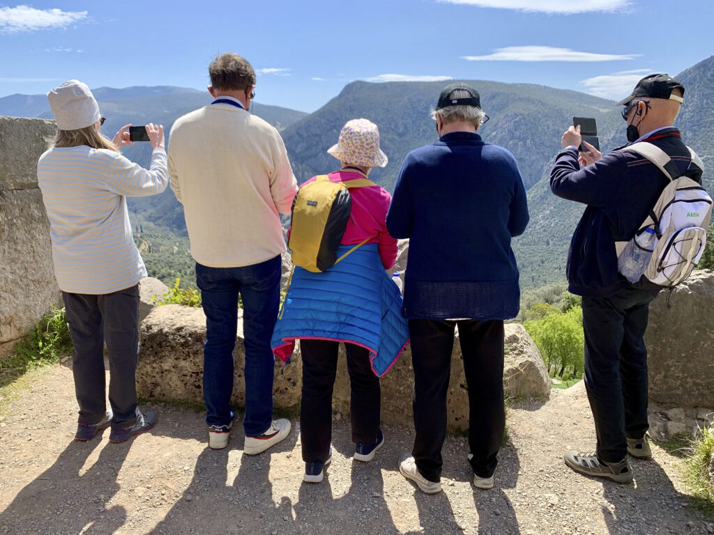 Fem personer står sida vid sida på en utsiktsplats och tittar bort och tar bilder av bergslandskapet. De bär jackor och hattar och en person har en ryggsäck. Vädret ser ut att vara soligt och klart.