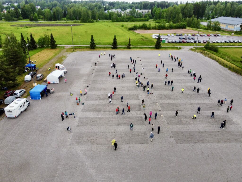 Flygfoto av människor utspridda i små grupper över en stor, öppen grusplan med träd och parkerade bilar i bakgrunden. Flera tält och fordon är uppställda på ena sidan av tomten.
