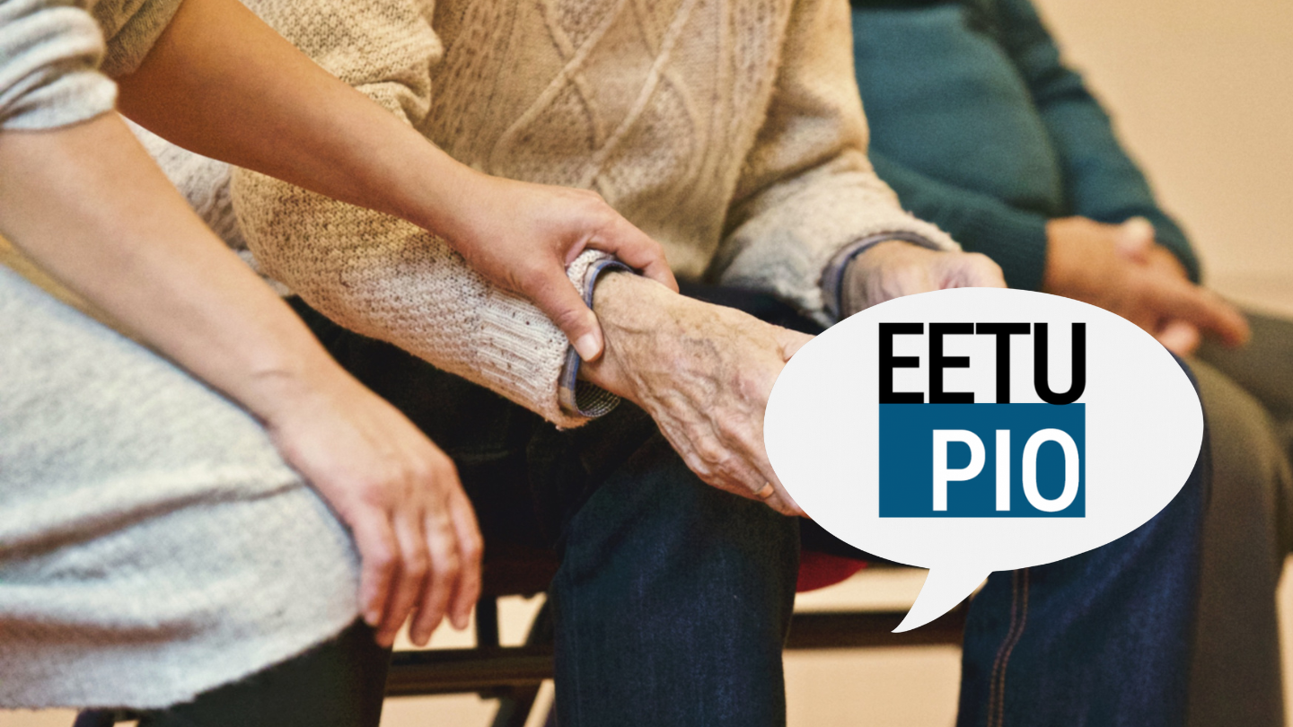 En person lägger en tröstande hand på armen på en äldre vuxen person i stickad tröja, med en annan sittande person synlig i bakgrunden. I en pratbubbla står det "EETU PIO.".