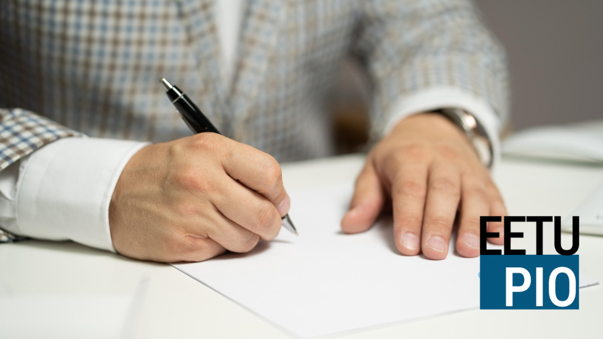 En person i rutig kavaj skriver på ett vitt papper med en svart penna. Texten EETU PIO visas i bildens nedre högra hörn.