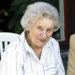 En äldre kvinna med kort, vågigt grått hår sitter på en vit stol. Hon bär en vit blus med tunna blå ränder och runda örhängen. I bakgrunden syns gröna växter och ett fönster med röd ram.