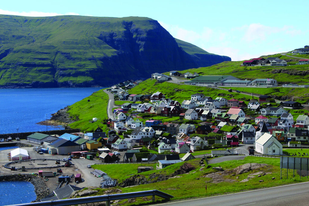 Liten kustby med färgglada hus på en grön sluttning vid havet, med ett brant berg i bakgrunden och några båtar vid vattnet. Himlen är mestadels klar.